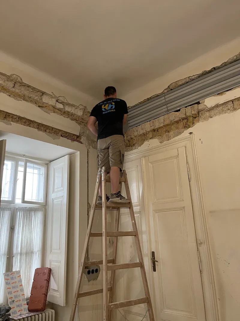Szerelő létrán védőcsöveket fektet a fal tetején kivésett horonyba